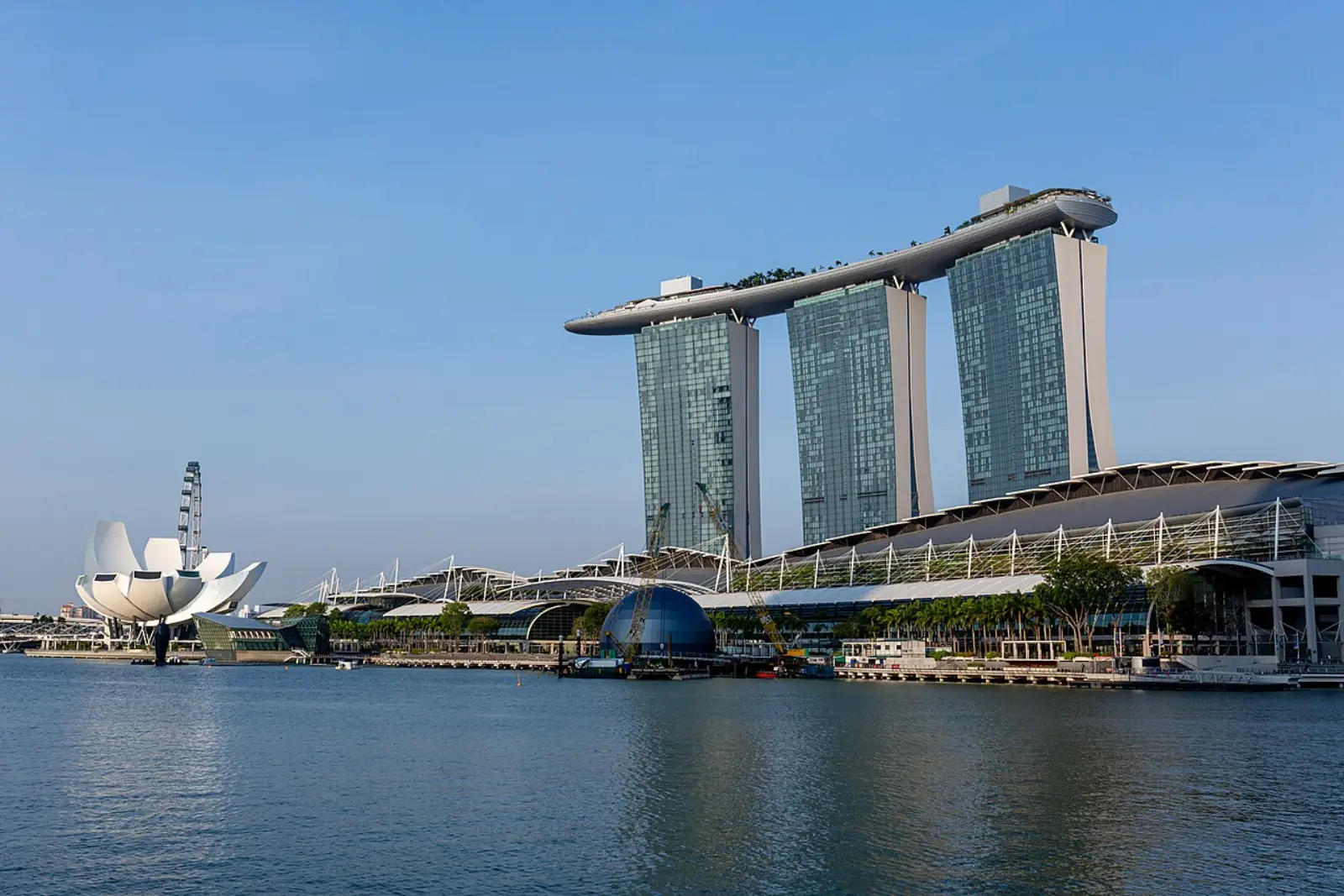 Marina Bay Sands seen across the water from the Singapore bayfront: three sloping towers carrying a single long deck, with the lotus-shaped ArtScience Museum and the convention centre roof to the left.