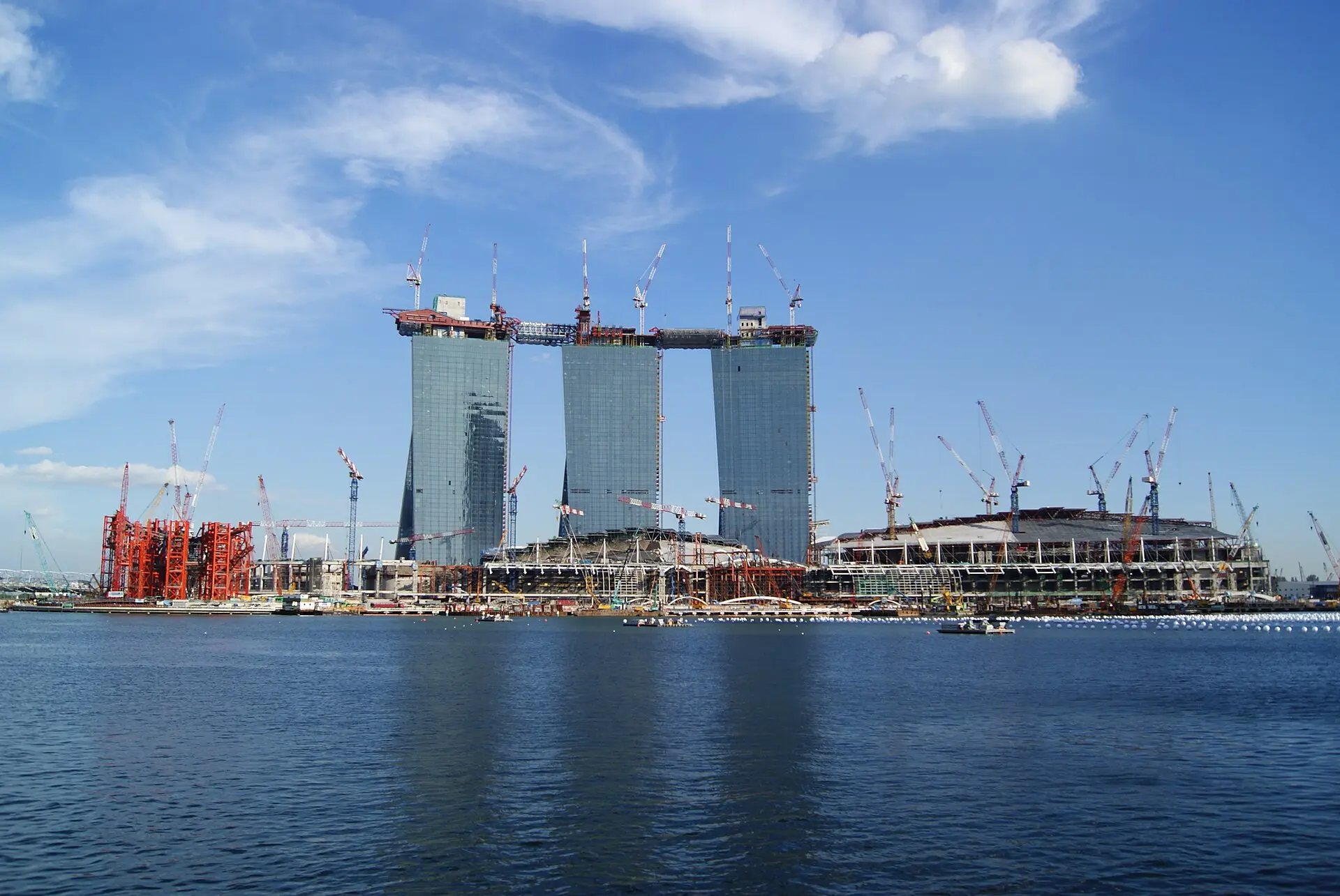 Marina Bay Sands under construction in November 2009 seen across the water: three bare concrete towers topped out with no deck yet spanning them, surrounded by dozens of tower cranes, with the steel frame of the convention centre and a red gantry to either side.