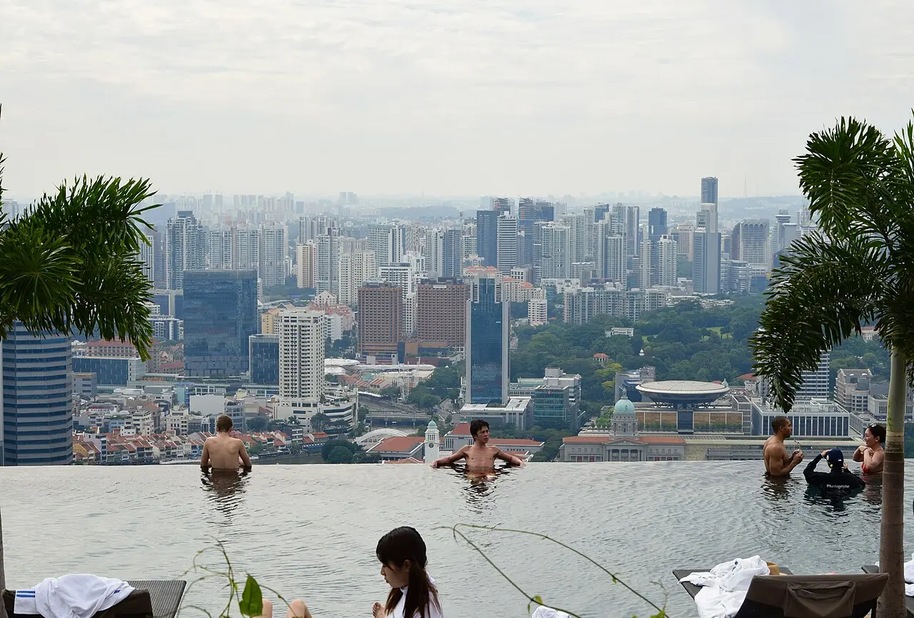 Swimmers standing at the far edge of the Marina Bay Sands infinity pool, looking out over the Singapore skyline in haze, with potted palms and a sun lounger with folded towels in the foreground.