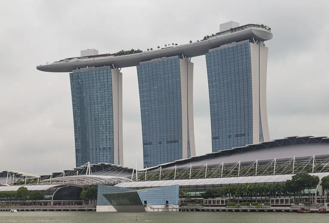 Close view of the three Marina Bay Sands towers under grey cloud, each tower splitting into two leaning legs, with the SkyPark deck running across their tops and projecting well past the left-hand tower into open air.