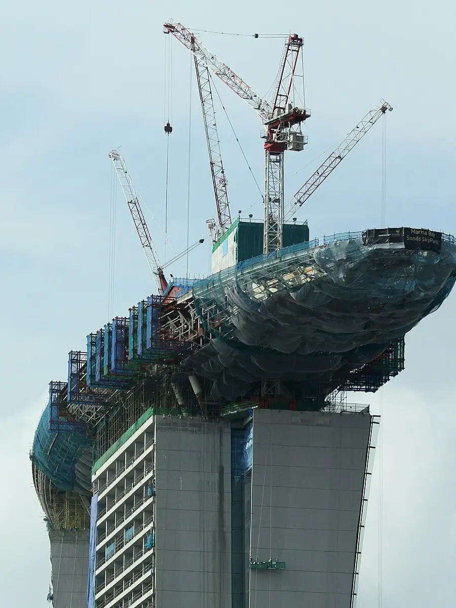 The head of one Marina Bay Sands tower in January 2010, still being built: the SkyPark deck under assembly on temporary steelwork, growing sideways out of the tower top with its far end hanging over open air, tower cranes standing above it and a Marina Bay Sands SkyPark banner on the deck edge.