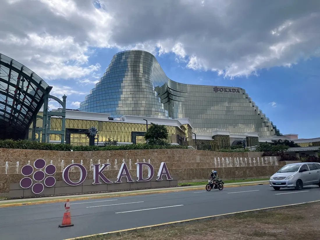 Okada Manila from the roadside: a long curved facade of gold and blue glass rising above a low stone wall, with a line of small fountain jets playing along the wall and a motorcycle and van passing on the road in front.