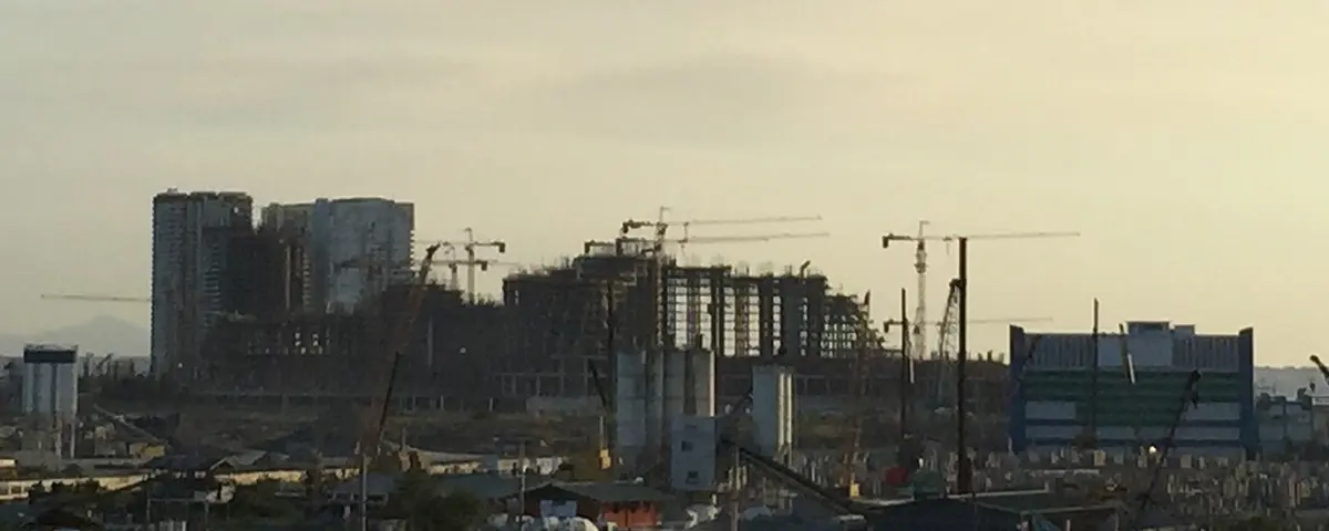 The Okada Manila site in April 2015 at dusk, then known as Manila Bay Resort: a long rectilinear concrete frame several storeys high standing under a row of tower cranes, with low-rise Parañaque in the foreground and the bay behind.