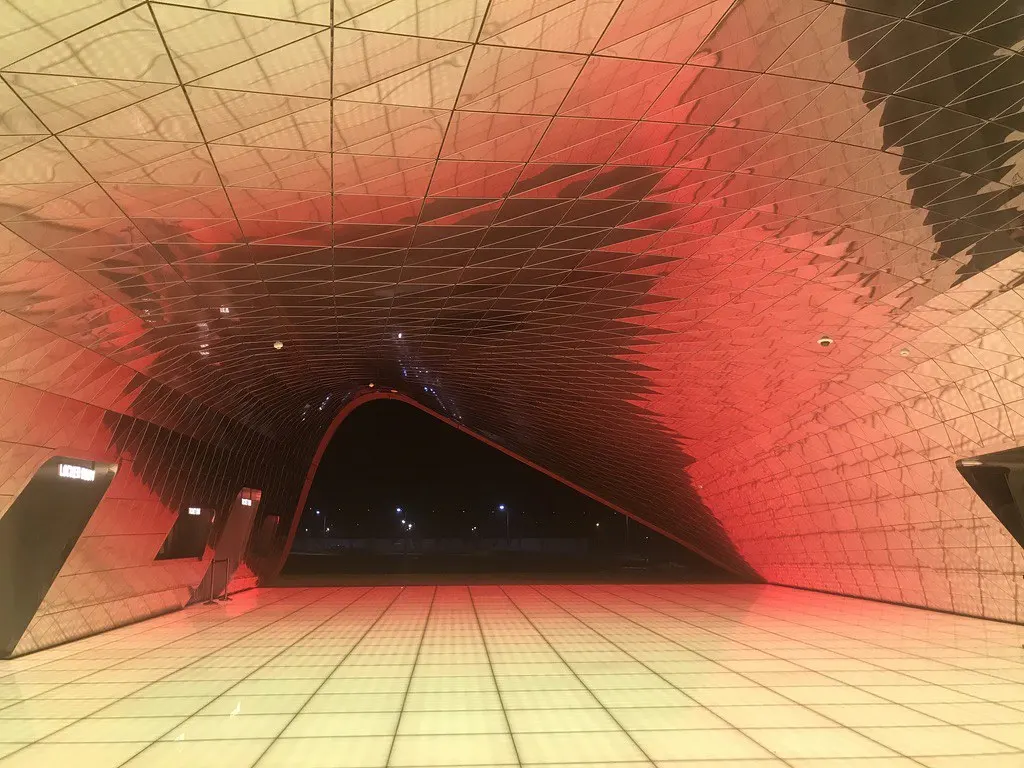 Interior of a doubly curved shell canopy at Paradise City, Incheon: a gridded funnel-shaped vault washed in red and amber light, narrowing to a dark opening onto the night outside, with a tiled floor running out through it.