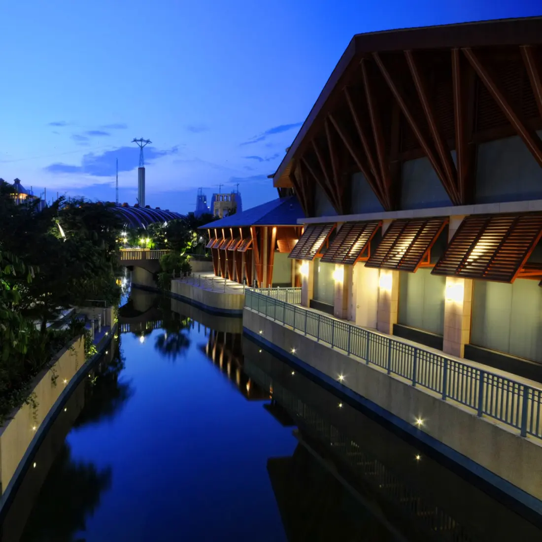 Resorts World Sentosa at dusk: a still canal running between low buildings, with a deep timber roof structure of exposed rafters and angled shading louvres lit from beneath on the right bank.