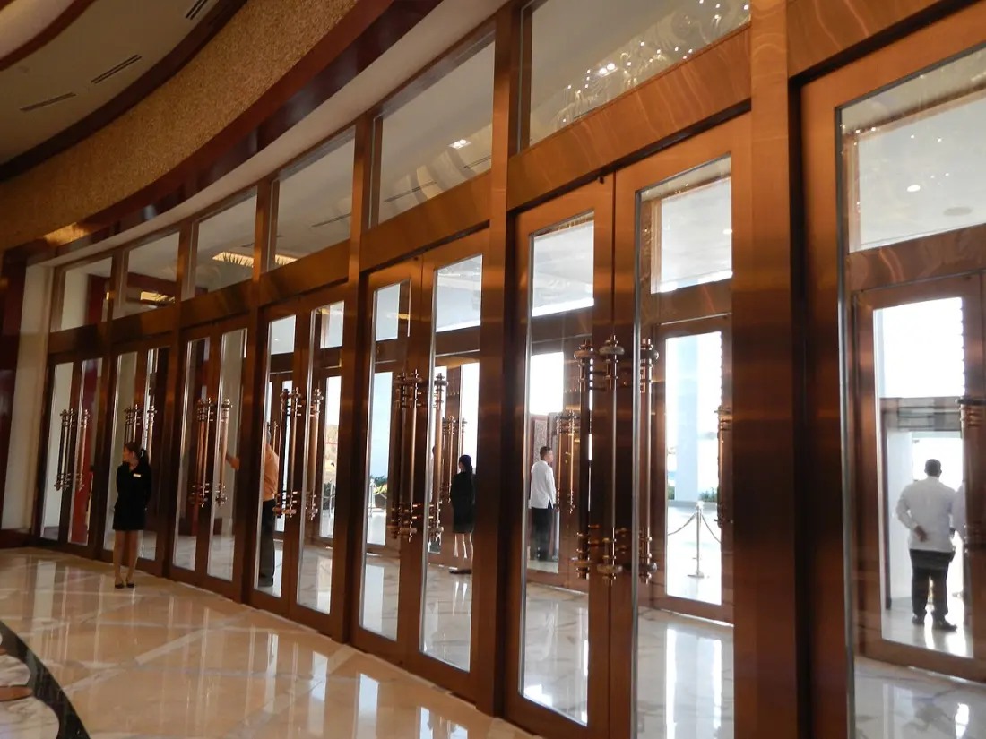 The entrance hall at Solaire Resort in Manila: a long shallow curve of tall bronze-framed glass doors with heavy vertical handles, polished marble floor, and staff and guests standing at the doors for scale.